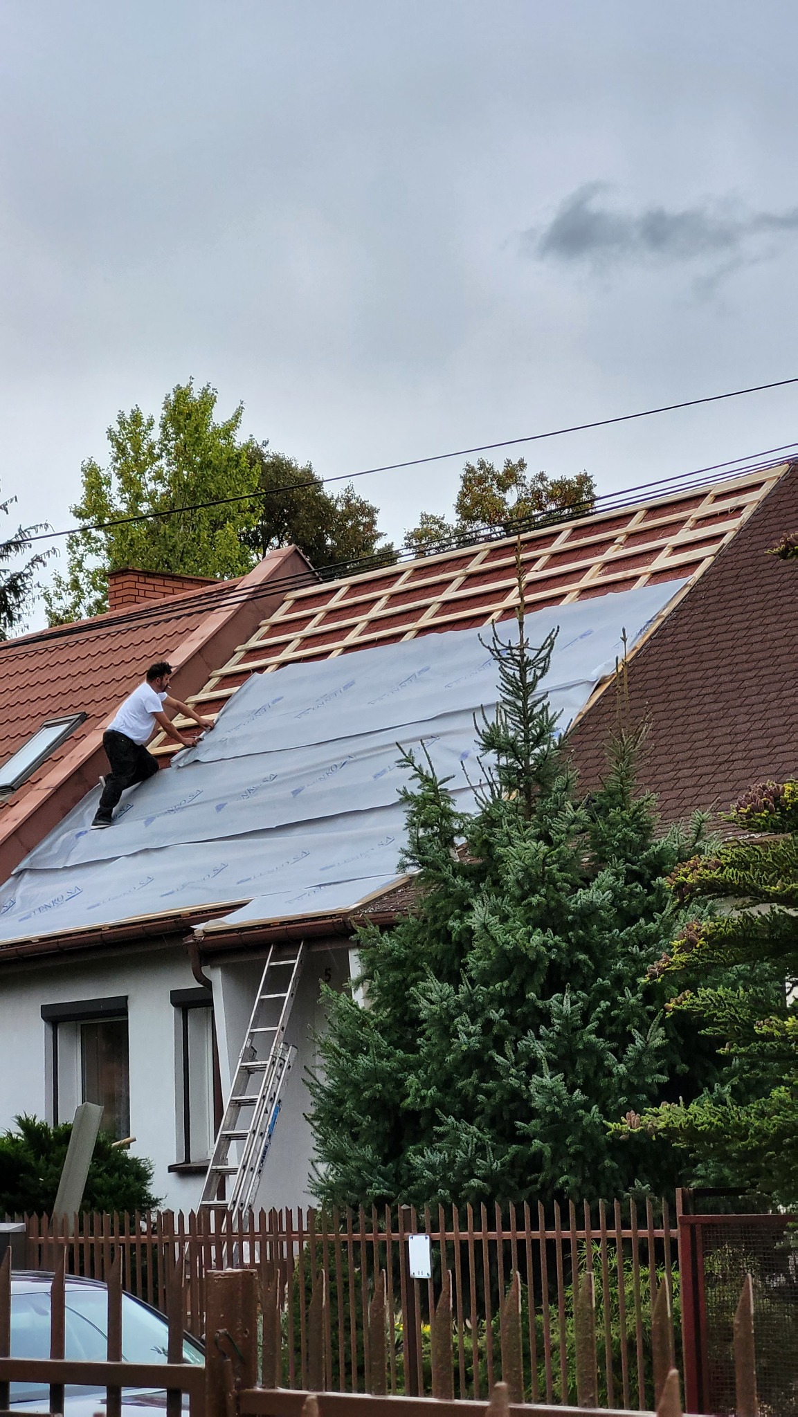 Pracownik w białej koszulce układa membranę dachową na drewnianej konstrukcji dachu domu jednorodzinnego z brązową dachówką, widok z poziomu gruntu, drabina oparta o ścianę budynku, pochmurne niebo.