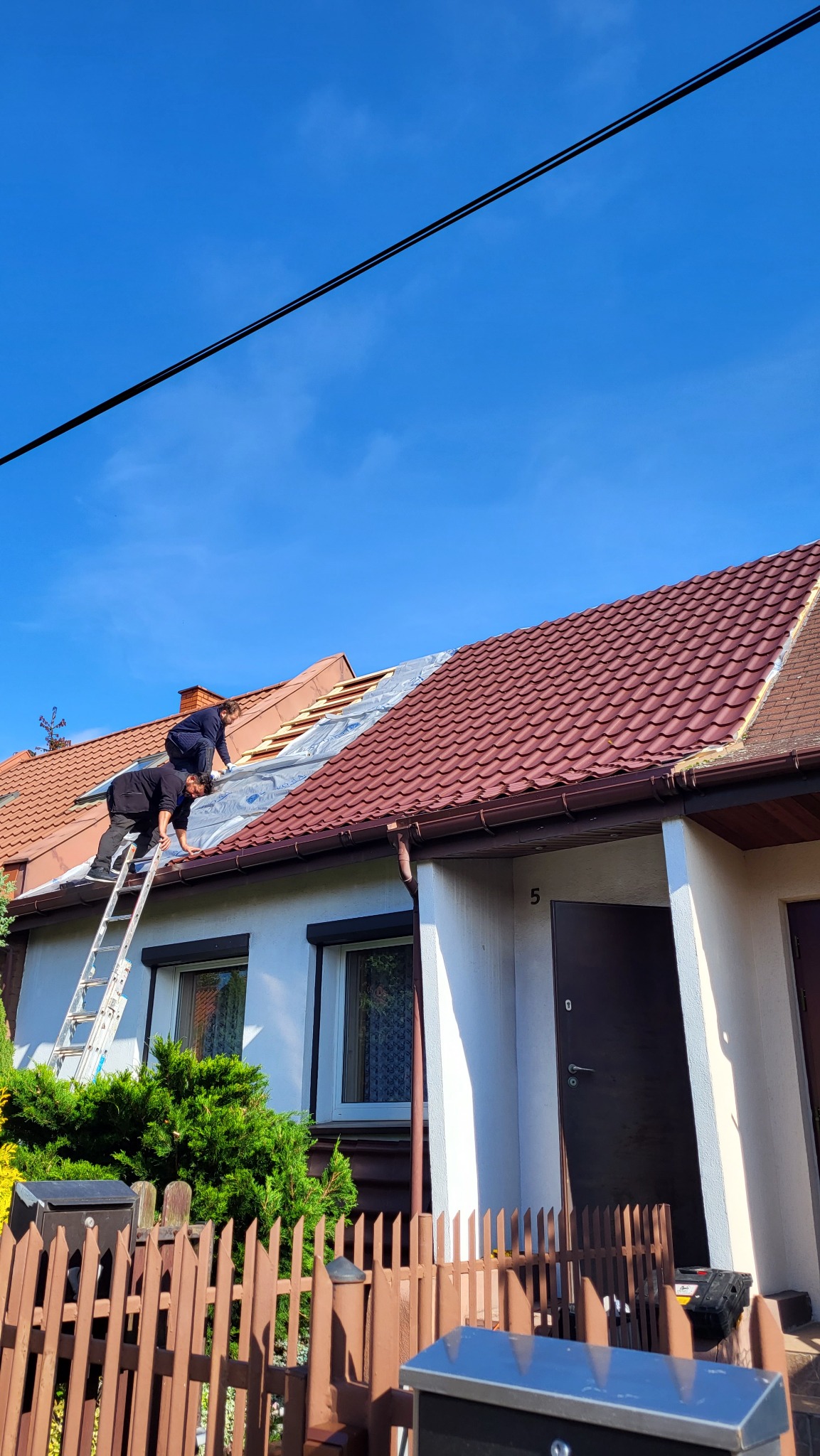 Dwóch dekarzy na drabinie układa membranę na dachu domu z czerwoną dachówką, widok na błękitne niebo.
