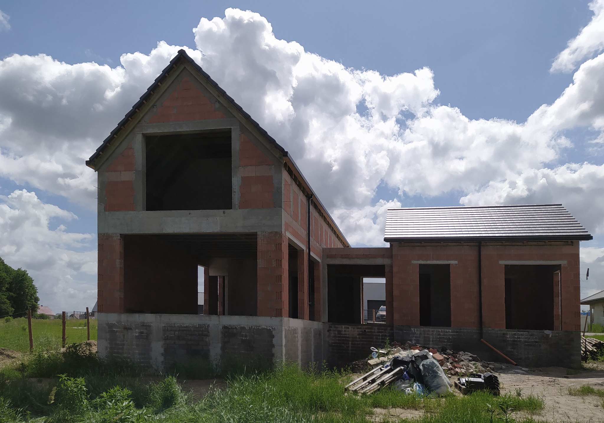 Surowy stan budynku jednorodzinnego z czerwonej cegły pod błękitnym niebem z obłokami, widoczne otwory okienne i drzwiowe, częściowo zadaszony, z materiałami budowlanymi na pierwszym planie.