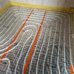 Rozłożona instalacja ogrzewania podłogowego z białych rurek na folii aluminiowej z czarną kratką, przymocowana plastikowymi klipsami, z pomarańczowymi pasami grzewczymi, widoczne ściany z białego...