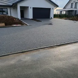 Nowo ułożona, szara kostka brukowa na podjeździe do nowoczesnego domu jednorodzinnego z garażem; widoczne kable i niewielka pryzma ziemi obok domu.