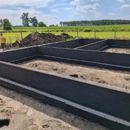 Wylane, pomalowane na czarno fundamenty domu jednorodzinnego z widocznymi zbrojeniami, na tle pola uprawnego i błękitnego nieba z obłokami.