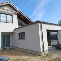 Świeżo otynkowany dom jednorodzinny z szarą stolarką okienną i drzwiami wejściowymi, z widocznymi śladami prac wykończeniowych wokół wejścia i samochodem dostawczym zaparkowanym obok garażu.