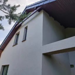 Jasna elewacja budynku z ciemną drewnianą podbitką dachową, widok z dołu na narożnik budynku, fragment drzewa iglastego w kadrze.