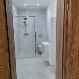 Nowoczesna, biała łazienka z marmurowym wzorem płytek, widziana zza drewnianych drzwi w Chorzelach. Kabina prysznicowa typu walk-in z deszczownicą, biała umywalka z szafką i wisząca toaleta.