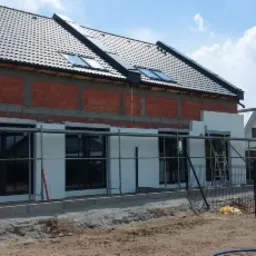 Dom jednorodzinny w trakcie ocieplania elewacji: fragment ściany z cegły, fragment z białym styropianem, rusztowanie z lewej strony, okna z czarnymi ramami, dach z dachówką w kolorze antracytowym...