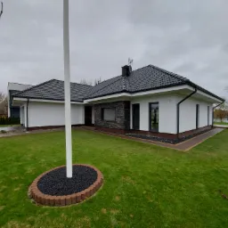 Nowoczesny parterowy dom z białymi ścianami, czarnym dachem z dachówki ceramicznej, kamienną okładziną elewacyjną i starannie utrzymanym trawnikiem w pochmurny dzień.