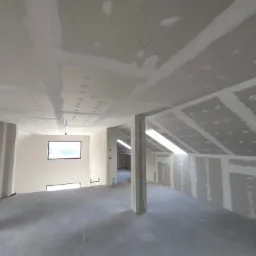 Poddasze w trakcie wykańczania: surowe ściany z płyt gipsowo-kartonowych, widoczne łączenia szpachlowane, betonowa wylewka podłogowa, okna dachowe i pionowy słup konstrukcyjny.