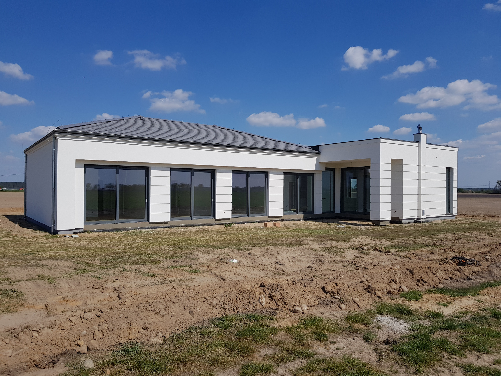 Nowoczesny parterowy dom z białymi ścianami i dużymi oknami na tle błękitnego nieba z chmurami, teren wokół w trakcie prac ziemnych.