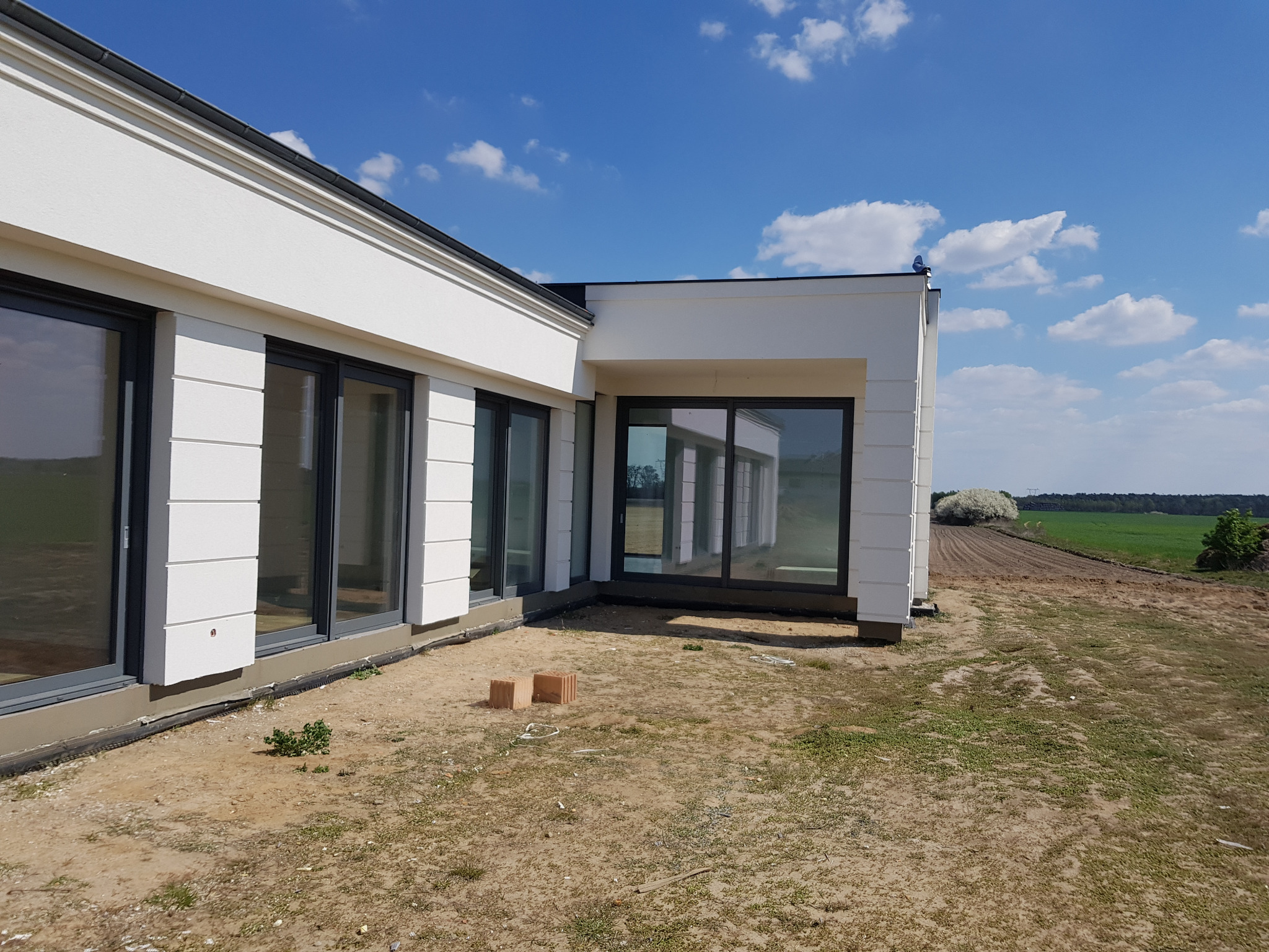 Nowoczesny dom jednorodzinny z białą elewacją, dużymi oknami i widokiem na pole uprawne w słoneczny dzień. Dwa czerwone cegły leżą na ziemi przed budynkiem.