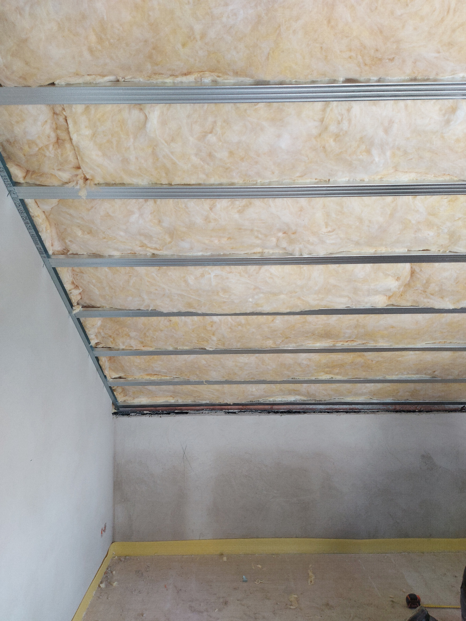 Poddasze w trakcie ocieplania: wełna mineralna umieszczona między metalowymi profilami sufitowymi, widoczne fragmenty ścian w stanie surowym i zabezpieczenia podłogi.