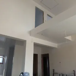 Wnętrze w trakcie prac wykończeniowych, widoczne ściany po nałożeniu gładzi, przygotowane do malowania, z widocznymi otworami drzwiowymi i schodami na antresolę.