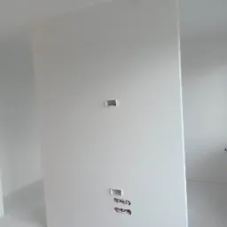 Wnętrze w trakcie wykańczania: świeżo pomalowane na biało ściany z widocznymi puszkami elektrycznymi i przygotowanymi otworami na gniazdka, jasne światło dzienne.