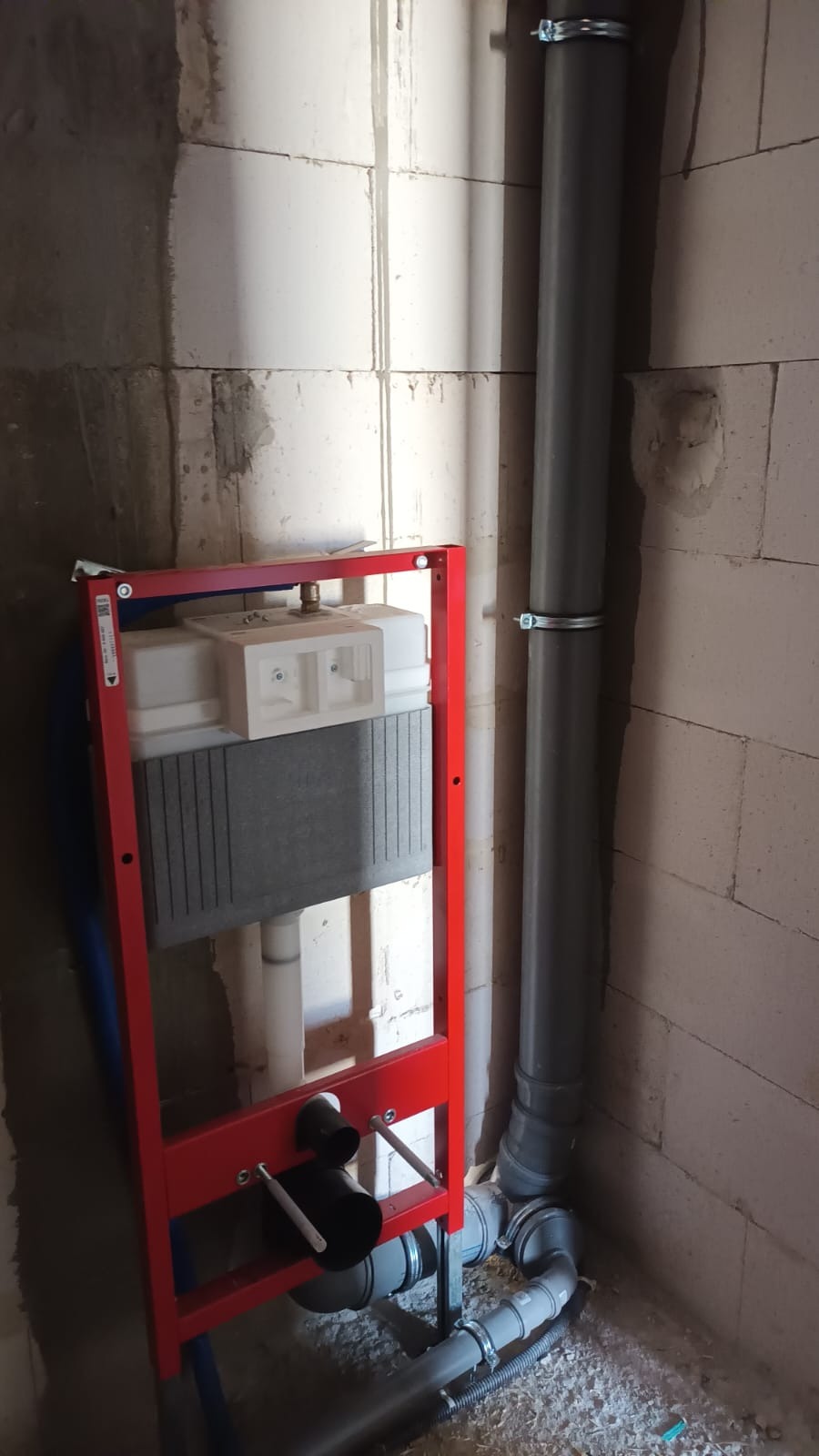 Stelaż podtynkowy WC w czerwonej ramie, widoczne szare rury kanalizacyjne przymocowane do ściany z betonu komórkowego, surowy stan wykończenia.