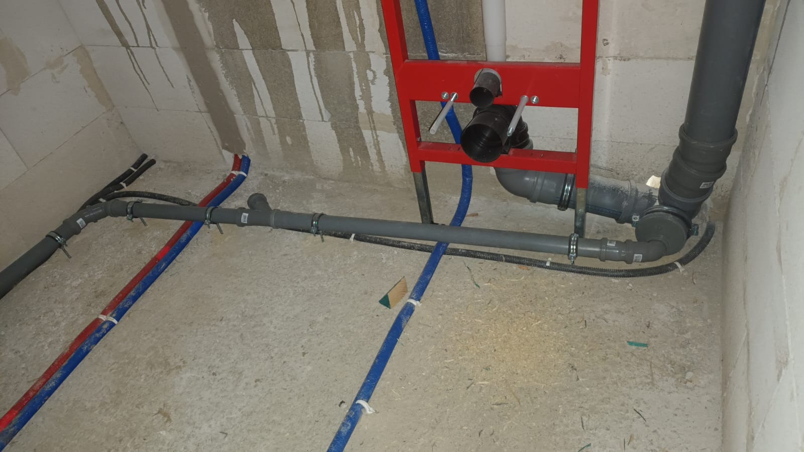 Surowe wnętrze z widoczną instalacją wodno-kanalizacyjną: szare rury kanalizacyjne, czerwone i niebieskie rury wodne, oraz czerwona rama montażowa podtynkowego systemu WC.