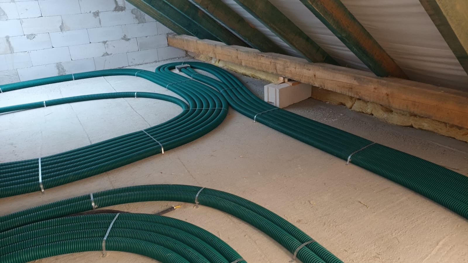 Zielone rury rekuperacji ułożone równolegle na poddaszu, zabezpieczone metalowymi obejmami, widoczne krokwie i izolacja.