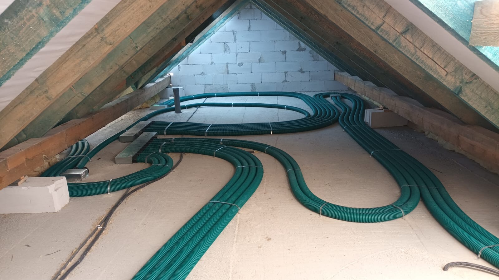 Zielone, karbowane rury wentylacyjne ułożone na poddaszu z drewnianą konstrukcją dachu i ścianą z białych bloczków.