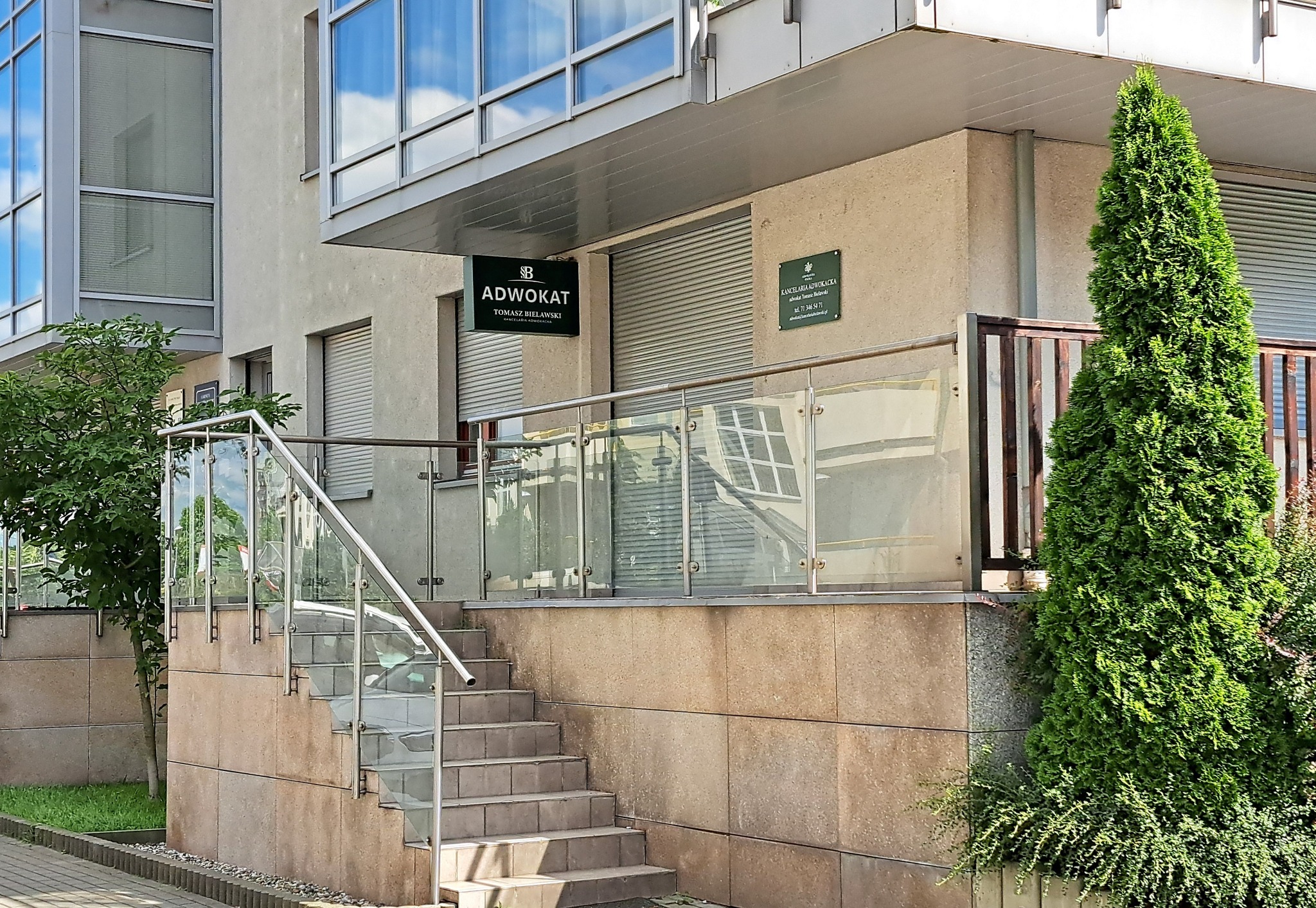 Nowoczesne wejście do kancelarii adwokackiej z beżową elewacją, szklaną balustradą przy schodach i zielonymi tujami. Widoczne szyldy 'ADWOKAT' i 'KANCELARIA ADWOKACKA'.