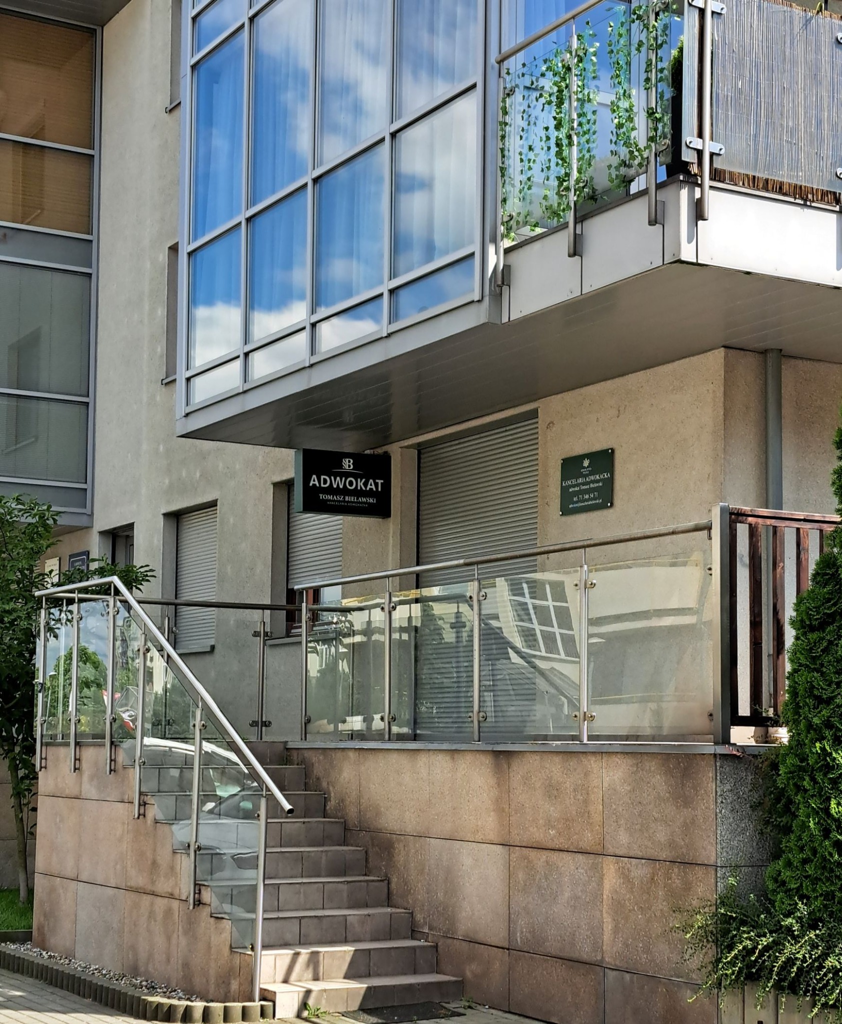 Kancelaria Adwokacka Tomasza Bielawskiego w nowoczesnym budynku z wejściem po schodach z przeszkloną balustradą, dużymi oknami odbijającymi niebo i zielenią.