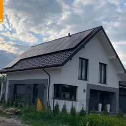 Nowoczesny dom jednorodzinny z panelami słonecznymi na dachu, widok z poziomu gruntu, pochmurne niebo w tle, ogrodzenie z siatki.