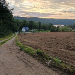 PPHU ECO-WARM BEATA SZLACHCIKOWSKA - Przygotowane pole po usunięciu drzew, widok na dom i jezioro w oddali, ślady opon na piaszczystej drodze.