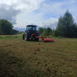 Ciemny traktor z pomarańczową kosiarką bijakową na polu po koszeniu, widok z tyłu, drzewa w tle, pochmurne niebo.