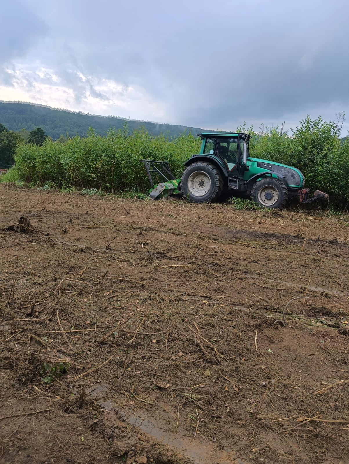 Zielony traktor Valtra z zamontowanym mulczerem do gałęzi na świeżo wykarczowanym terenie z widocznymi pniami i gałęziami, w tle zalesione wzgórza pod pochmurnym niebem.