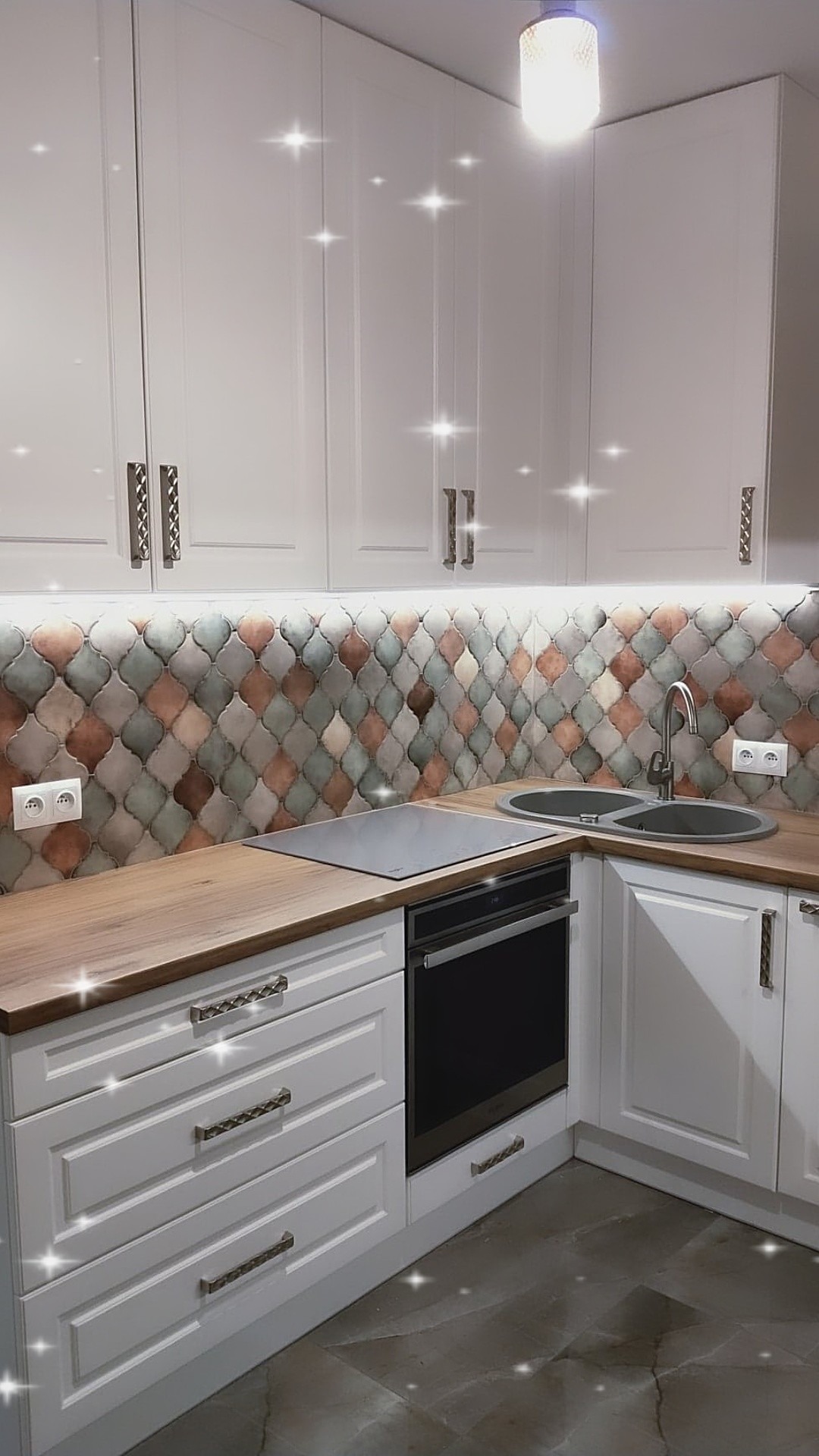 Jasna kuchnia z białymi szafkami, drewnianym blatem i dekoracyjną mozaiką na ścianie nad blatem, oświetlona listwą LED.