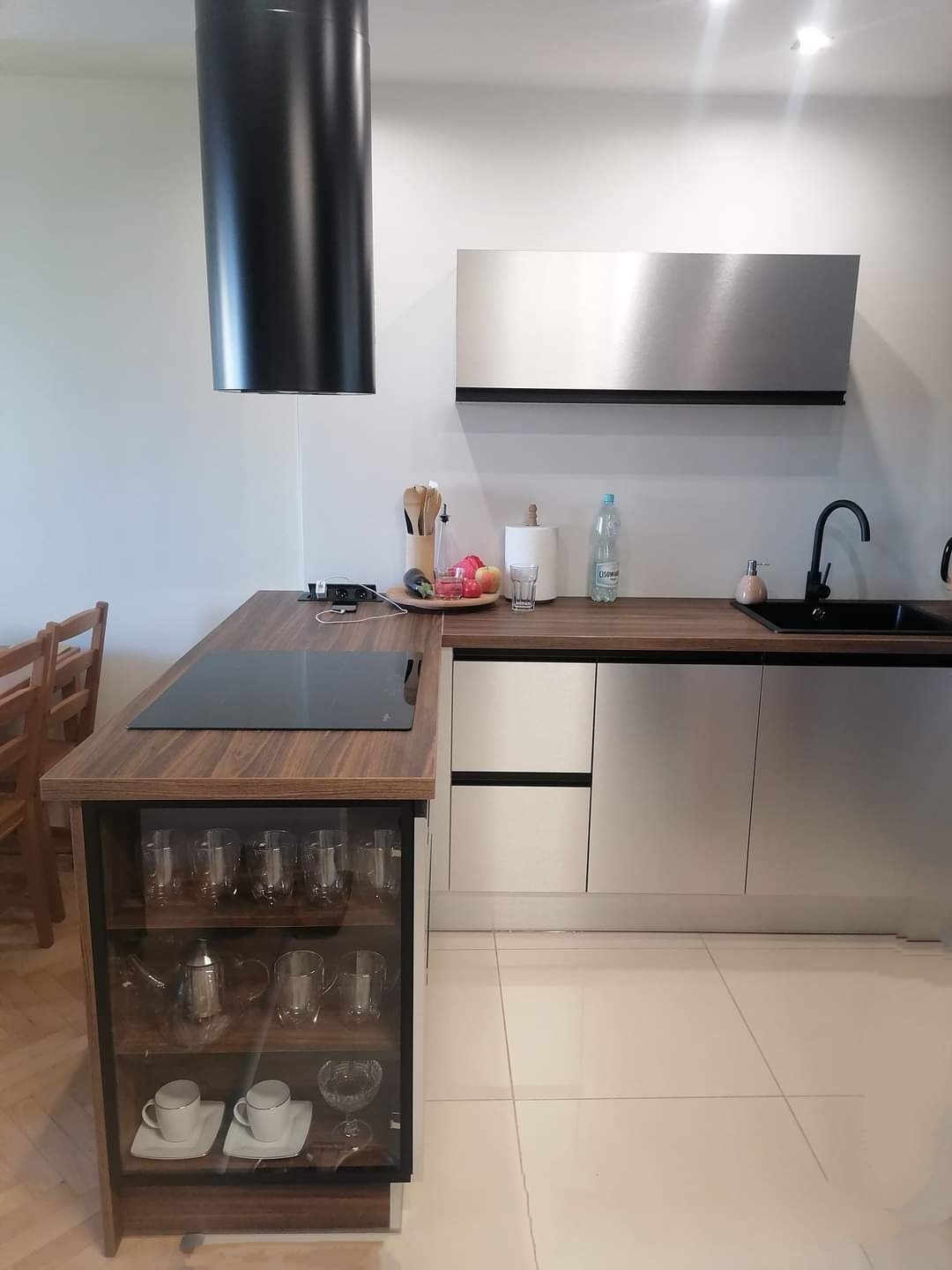 Nowoczesna kuchnia z drewnianym blatem, szarymi frontami szafek i przeszkloną witryną z zastawą, czarny okap cylindryczny.