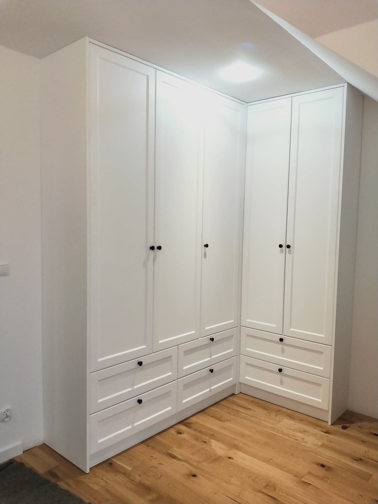 Biała szafa wnękowa z czarnymi gałkami, zabudowa narożna z szufladami, na podłodze z jasnego drewna.