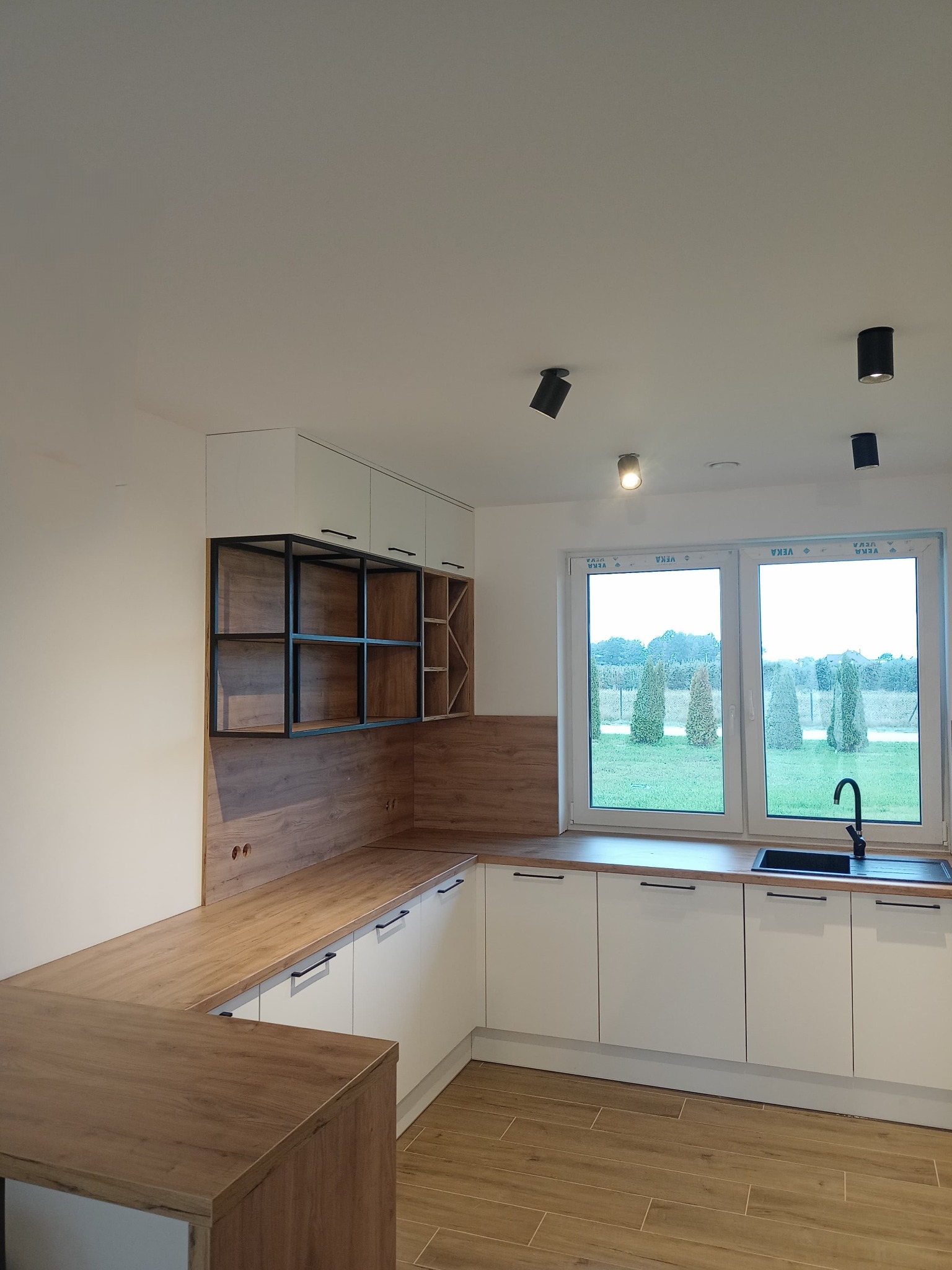 Nowoczesna kuchnia z białymi szafkami, drewnianym blatem i czarnymi akcentami, takimi jak armatura i metalowy regał nad blatem, oświetlona reflektorami sufitowymi, z widokiem na ogród przez okno.