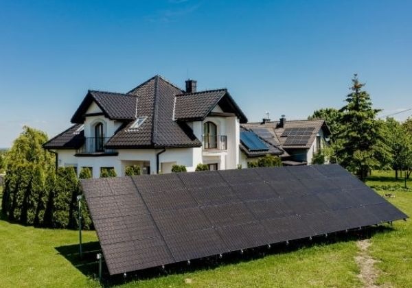 Czarny panel fotowoltaiczny na trawie przed domem z panelami na dachu, w tle błękitne niebo.