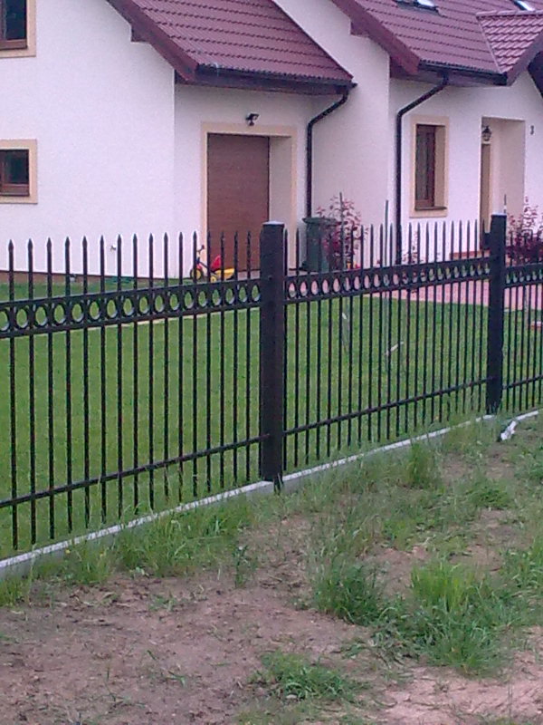 Czarny metalowy płot z elementami dekoracyjnymi w postaci okręgów, oddzielający trawnik od domu z beżową elewacją i brązowym dachem.