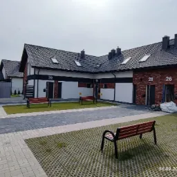 Nowe budynki szeregowe z numerami 26, 28, 30 i 32, wykończone cegłą klinkierową i białym tynkiem, z czarnymi dachami i oknami dachowymi, otoczone trawnikiem i brukowanym chodnikiem, z ławkami...