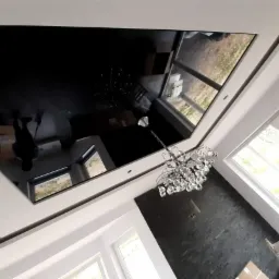 Błyszczący czarny sufit napinany z odbiciem pomieszczenia, widoczny żyrandol kryształowy i drabina oparta o ścianę z cegły.