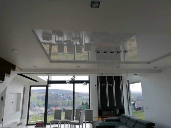 Nowoczesny salon z dużymi oknami i lśniącym, białym sufitem podwieszanym, w którym odbija się część wyposażenia, a nad nim dodatkowe oświetlenie punktowe i wiszące lampy.