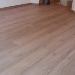 Ułożona podłoga z płytek imitujących drewno w odcieniach beżu, z widocznymi fugami i listwami przypodłogowymi. W tle fragment ściany w trakcie prac wykończeniowych.