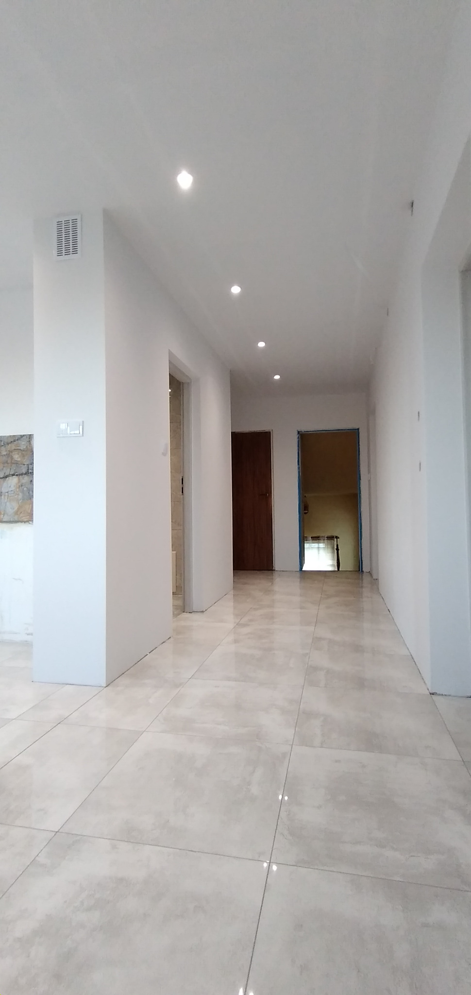 Jasne płytki podłogowe w korytarzu z białymi ścianami i sufitami z punktowym oświetleniem, widoczne drzwi do łazienki i pokoju.