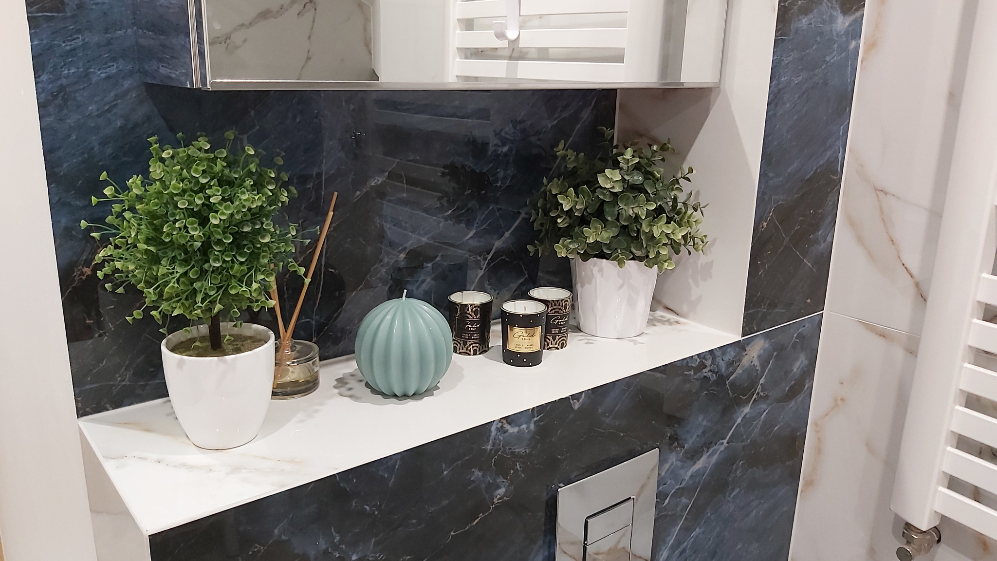 Półka w łazience z dekoracjami: dwie rośliny w białych doniczkach, świeca w kształcie dyni i trzy małe świeczki zapachowe na tle płytek imitujących marmur w odcieniach bieli i granatu.