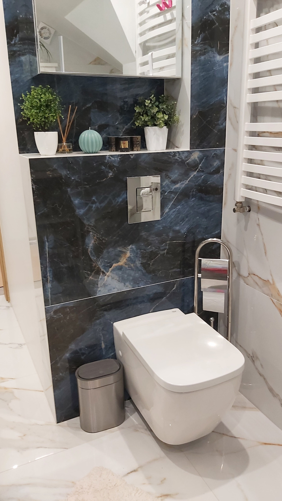 Nowoczesna toaleta podwieszana na tle ściany z płytek imitujących marmur z dekoracyjnymi akcentami roślinnymi i grzejnikiem drabinkowym.