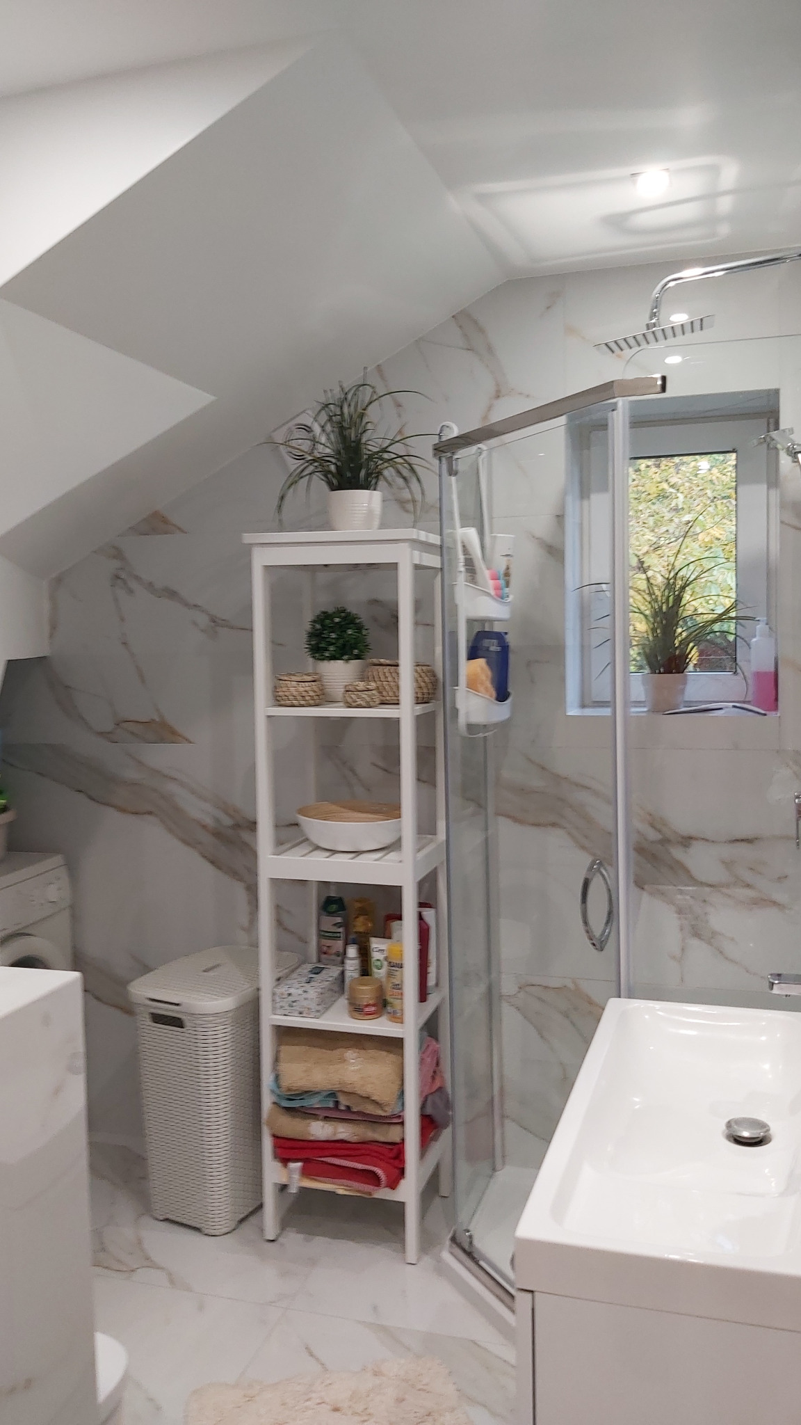 Jasna łazienka na poddaszu z marmurowymi płytkami, kabiną prysznicową, białą szafką regałową z dekoracjami i roślinami oraz pralką w tle.
