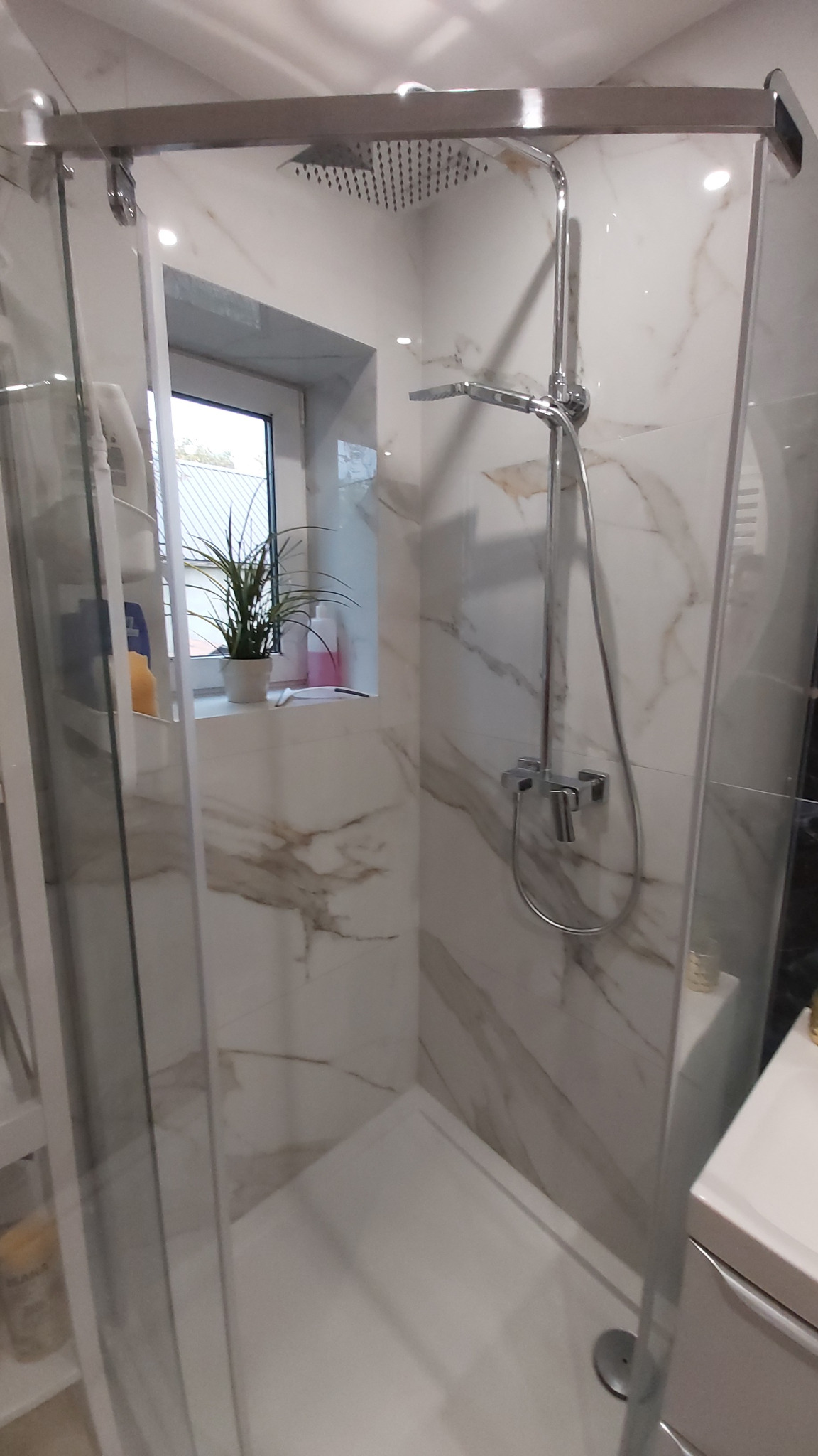 Nowoczesna kabina prysznicowa z deszczownicą i ręcznym prysznicem, wykończona płytkami imitującymi marmur z subtelnym oknem z rośliną w tle.