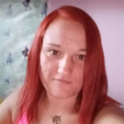 Portret kobiety z rudymi włosami w różowym topie, na tle ściany z motywami z filmu 'Kraina Lodu', widoczna półka z kosmetykami.