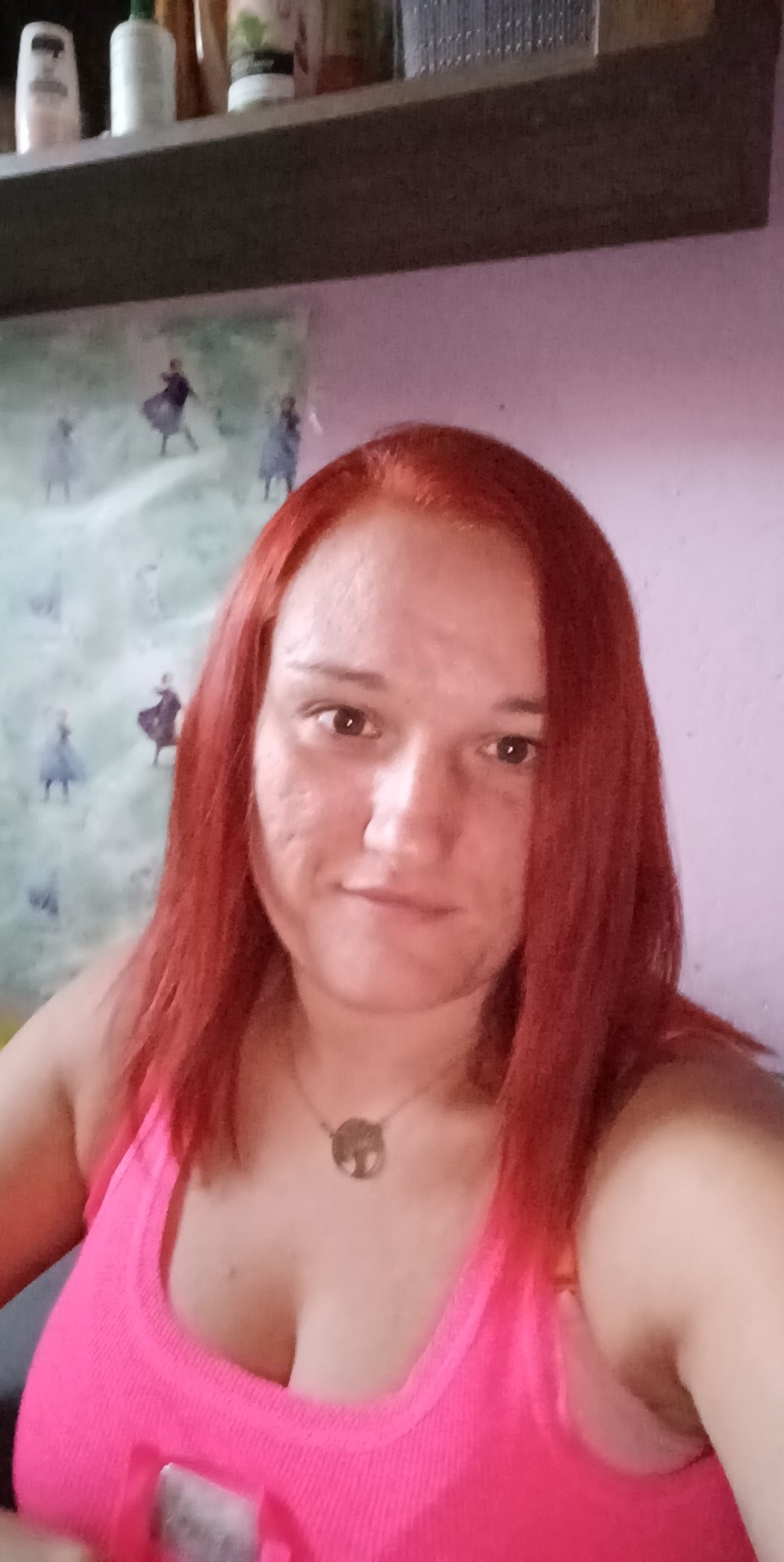 Portret kobiety z rudymi włosami w różowym topie, na tle ściany z motywami z filmu 'Kraina Lodu', widoczna półka z kosmetykami.