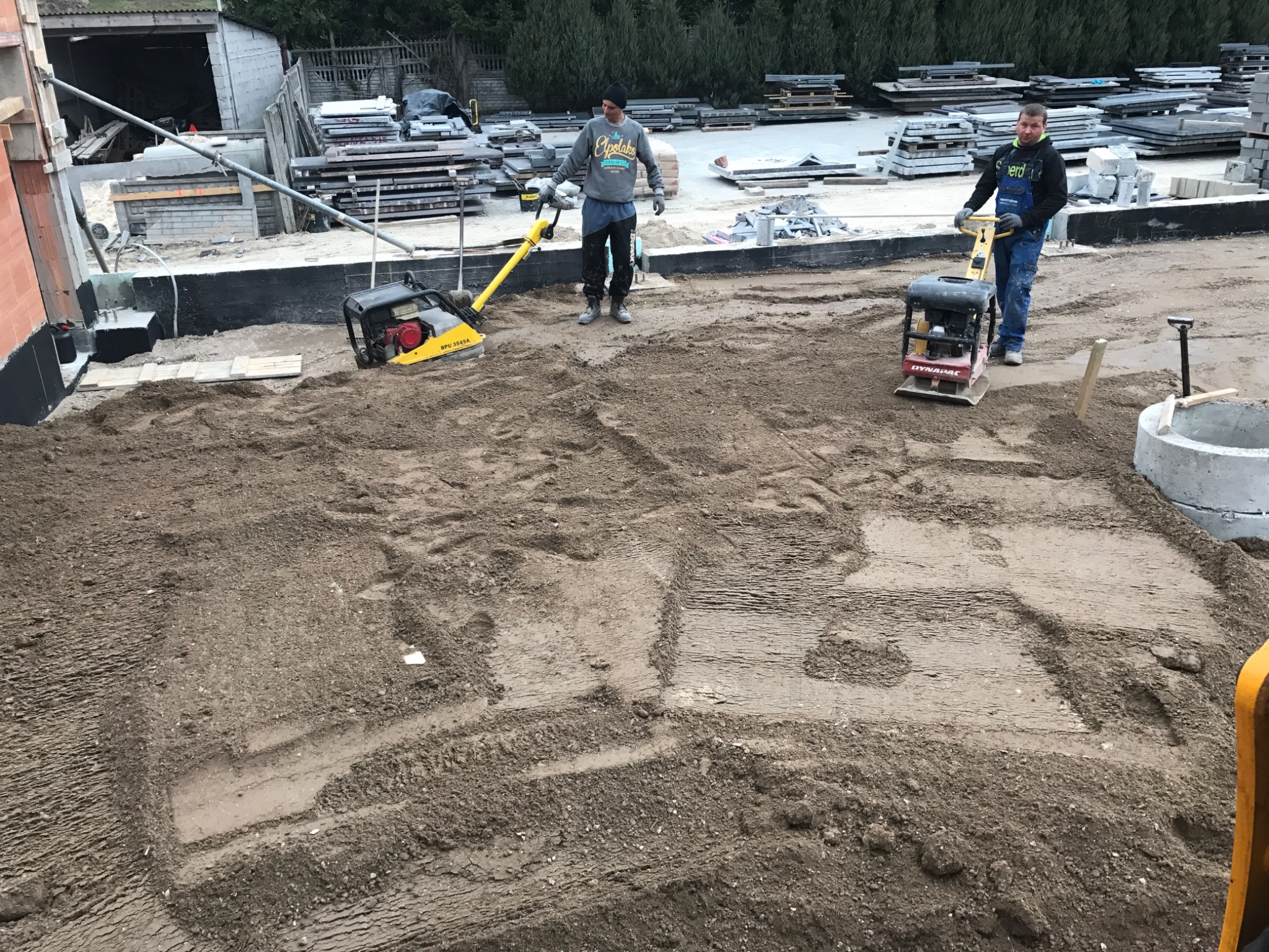 Dwóch pracowników zagęszcza piasek pod fundamenty za pomocą zagęszczarek wibracyjnych. Widoczne ślady po zagęszczaniu i przygotowany szalunek pod wylewkę.