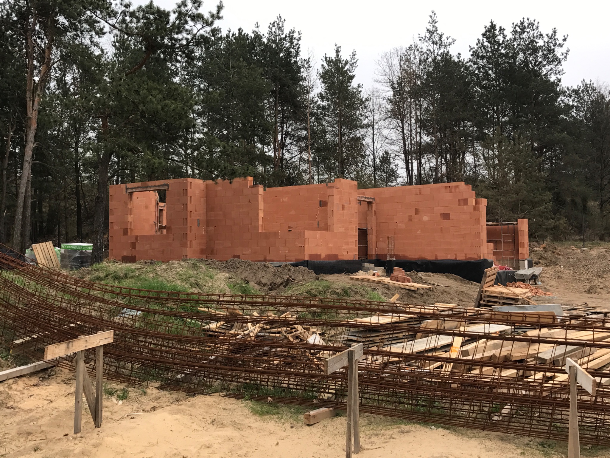 Budowa domu z czerwonej cegły, widoczne ściany w trakcie murowania, zbrojenie na pierwszym planie, teren budowy otoczony lasem.