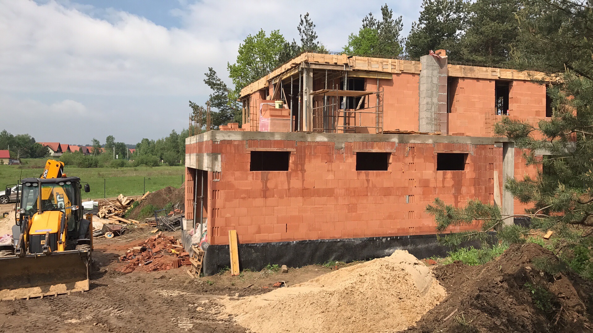 Budowa domu z czerwonej cegły ceramicznej, widoczny dwupoziomowy budynek w stanie surowym otwartym, z żółtą koparką w tle i stertami gruzu oraz piasku na pierwszym planie.