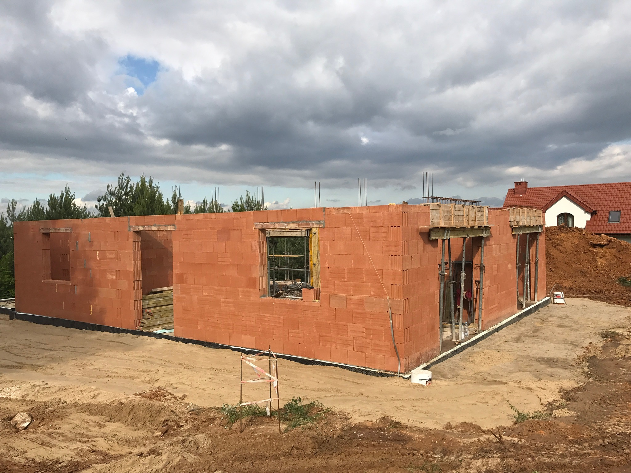 Surowy stan domu z czerwonej cegły, widoczne otwory okienne i drzwiowe, częściowo oszalowany strop, teren wokół w stanie budowy, w tle dom z czerwoną dachówką.
