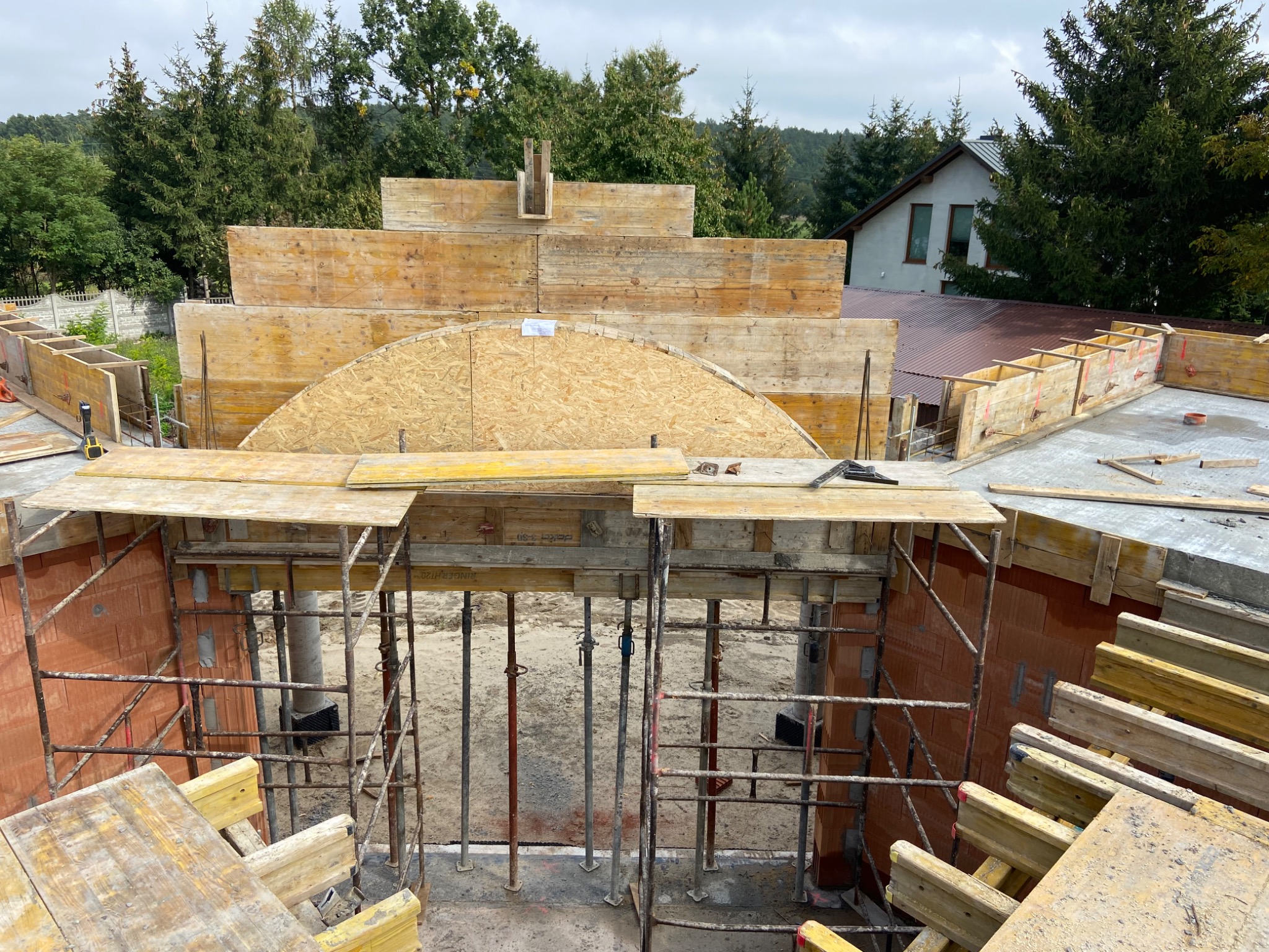 Wysoki kąt na budowę z drewnianymi szalunkami, stalowymi podporami i fragmentem muru z czerwonej cegły, z widokiem na drzewa i dom w tle.