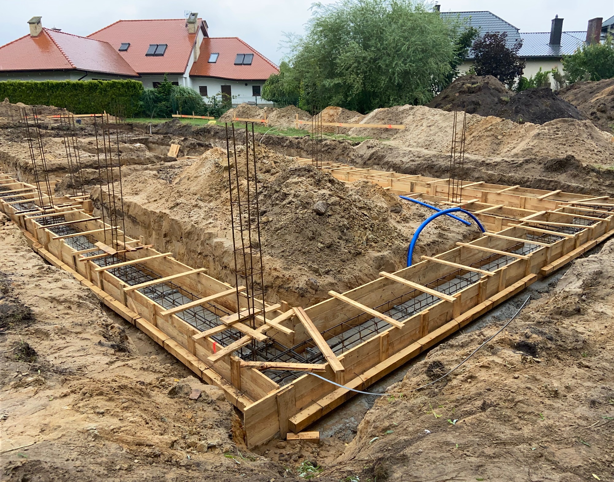 Przygotowanie do wylewki fundamentów: drewniany szalunek zbrojony stalą, widoczne niebieskie rury instalacyjne, w tle domy jednorodzinne i hałdy ziemi.